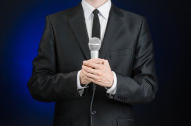 Business and speech topic: Man in black suit holding a microphone on a gray dark blue isolated background in studio