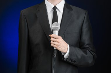Business and speech topic: Man in black suit holding a microphone on a gray dark blue isolated background in studio
