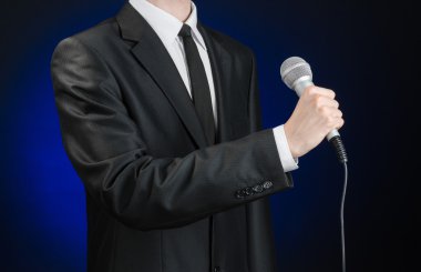 Business and speech topic: Man in black suit holding a microphone on a gray dark blue isolated background in studio