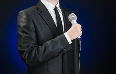 Business and speech topic: Man in black suit holding a microphone on a gray dark blue isolated background in studio