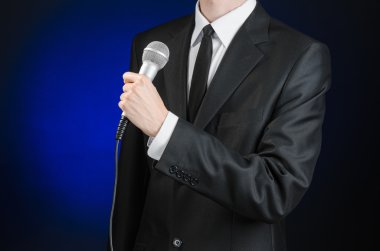 Business and speech topic: Man in black suit holding a microphone on a gray dark blue isolated background in studio
