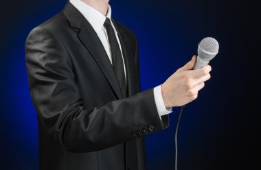 Business and speech topic: Man in black suit holding a microphone on a gray dark blue isolated background in studio
