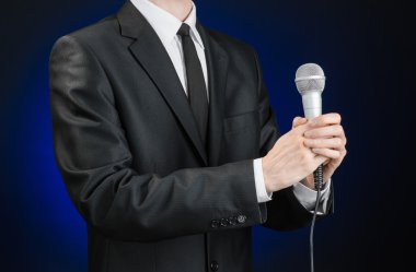 Business and speech topic: Man in black suit holding a microphone on a gray dark blue isolated background in studio