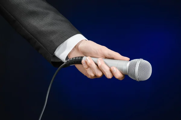 Business and speech topic: Man in black suit holding a microphone on a gray dark blue isolated background in studio — 图库照片