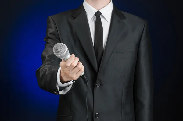 Business and speech topic: Man in black suit holding a microphone on a gray dark blue isolated background in studio — Stok fotoğraf