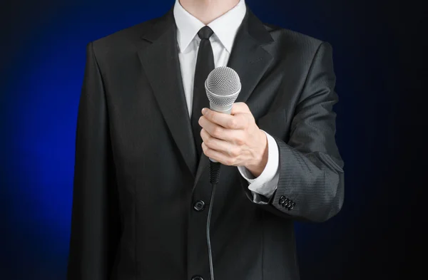 Business and speech topic: Man in black suit holding a microphone on a gray dark blue isolated background in studio — 图库照片