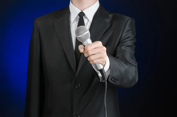 Business and speech topic: Man in black suit holding a microphone on a gray dark blue isolated background in studio — 스톡 사진