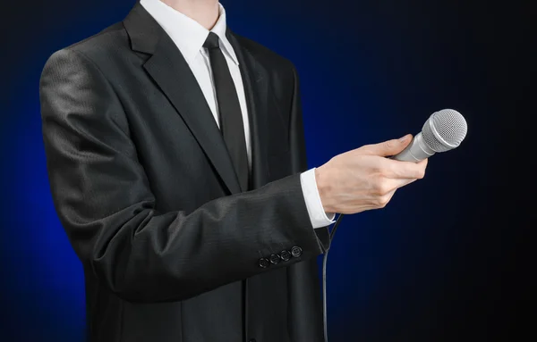 Business and speech topic: Man in black suit holding a microphone on a gray dark blue isolated background in studio — 스톡 사진