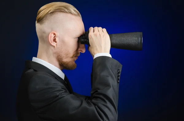 Téma podnikání a hledání: muž v černém obleku drží černý dalekohled v ruce na tmavě modrém pozadí ve studiu izolovaných — Stock fotografie