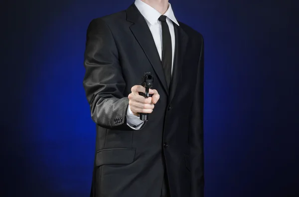 Armas de fuego y temas de seguridad: un hombre con un traje negro sosteniendo un arma sobre un fondo azul oscuro en un estudio aislado —  Fotos de Stock
