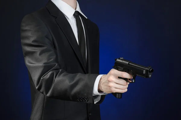 Schusswaffen und Sicherheitsthema: ein Mann im schwarzen Anzug mit Waffe auf dunkelblauem Hintergrund im Studio isoliert — Stockfoto