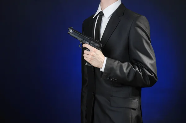 Armas de fogo e tópico de segurança: um homem de terno preto segurando uma arma em um fundo azul escuro em estúdio isolado — Fotografia de Stock