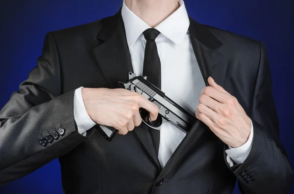 Зброя та тема безпеки: чоловік у чорному костюмі тримає пістолет на темно-синьому тлі в студії ізольований — стокове фото