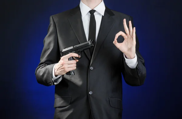 Armas de fuego y temas de seguridad: un hombre con un traje negro sosteniendo un arma sobre un fondo azul oscuro en un estudio aislado — Foto de Stock