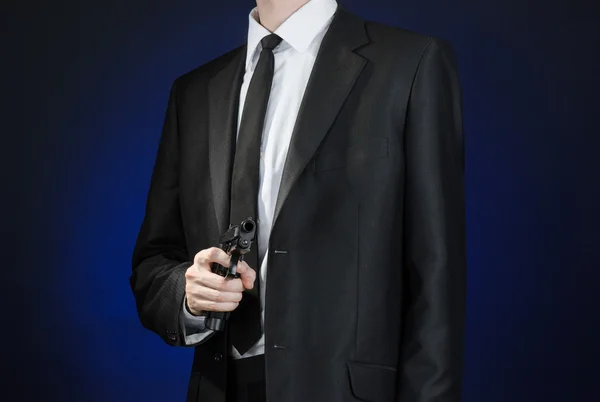 Armas de fuego y temas de seguridad: un hombre con un traje negro sosteniendo un arma sobre un fondo azul oscuro en un estudio aislado — Foto de Stock