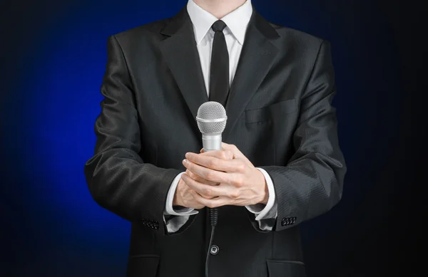 Business and speech topic: Man in black suit holding a microphone on a gray dark blue isolated background in studio Stock Photo