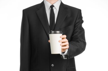 Business lunches coffee theme: businessman in a black suit holding a white blank paper cup of coffee with a brown plastic cap isolated on a white background in the studio, advertising coffee