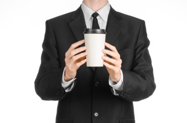 Business lunches coffee theme: businessman in a black suit holding a white blank paper cup of coffee with a brown plastic cap isolated on a white background in the studio, advertising coffee