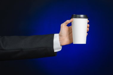 Business lunch and coffee theme: businessman in a black suit holding a white blank paper cup of coffee with a brown plastic cap on a dark blue background isolated in the studio, advertising coffee