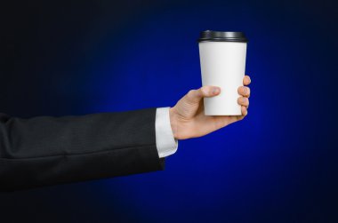 Business lunch and coffee theme: businessman in a black suit holding a white blank paper cup of coffee with a brown plastic cap on a dark blue background isolated in the studio, advertising coffee