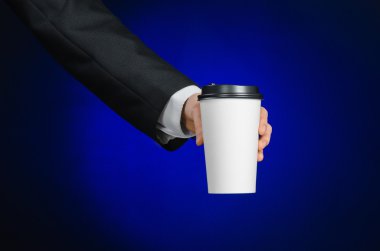 Business lunch and coffee theme: businessman in a black suit holding a white blank paper cup of coffee with a brown plastic cap on a dark blue background isolated in the studio, advertising coffee
