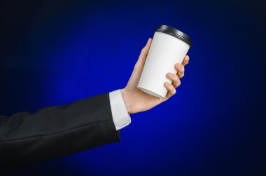 Business lunch and coffee theme: businessman in a black suit holding a white blank paper cup of coffee with a brown plastic cap on a dark blue background isolated in the studio, advertising coffee