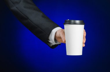 Business lunch and coffee theme: businessman in a black suit holding a white blank paper cup of coffee with a brown plastic cap on a dark blue background isolated in the studio, advertising coffee