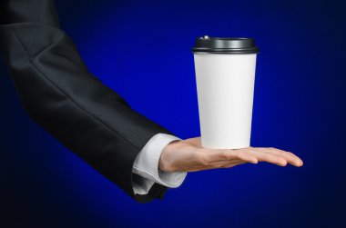 Business lunch and coffee theme: businessman in a black suit holding a white blank paper cup of coffee with a brown plastic cap on a dark blue background isolated in the studio, advertising coffee