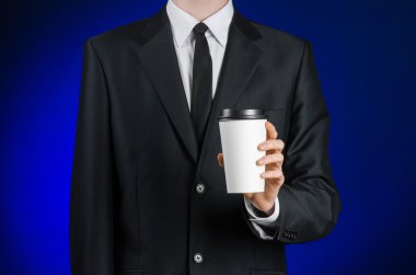 Business lunch and coffee theme: businessman in a black suit holding a white blank paper cup of coffee with a brown plastic cap on a dark blue background isolated in the studio, advertising coffee