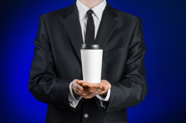 Business lunch and coffee theme: businessman in a black suit holding a white blank paper cup of coffee with a brown plastic cap on a dark blue background isolated in the studio, advertising coffee