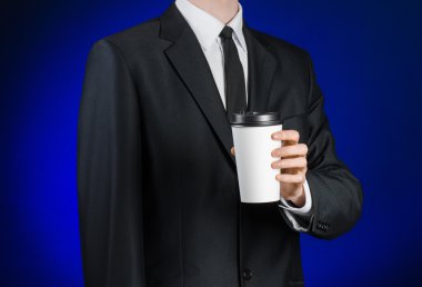 Business lunch and coffee theme: businessman in a black suit holding a white blank paper cup of coffee with a brown plastic cap on a dark blue background isolated in the studio, advertising coffee