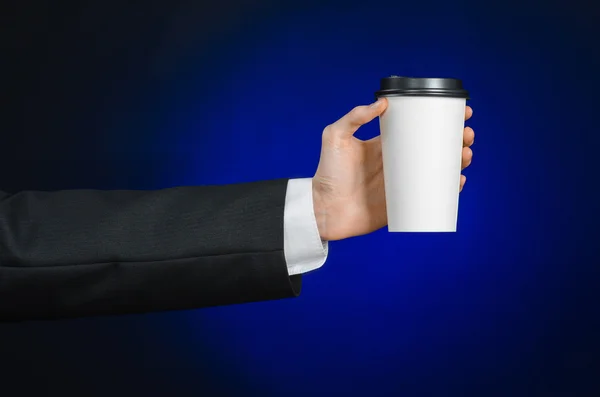 Business lunch and coffee theme: businessman in a black suit holding a white blank paper cup of coffee with a brown plastic cap on a dark blue background isolated in the studio, advertising coffee