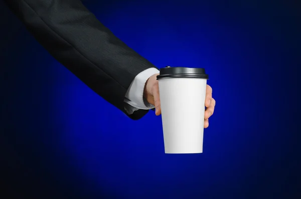 Business lunch and coffee theme: businessman in a black suit holding a white blank paper cup of coffee with a brown plastic cap on a dark blue background isolated in the studio, advertising coffee — ストック写真