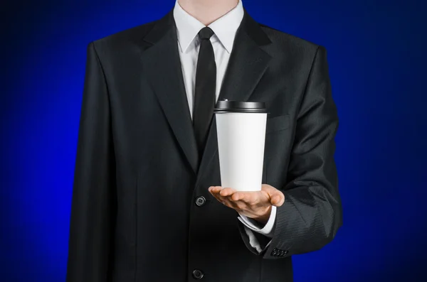 Almoço de negócios e tema de café: homem de negócios em um terno preto segurando uma xícara de café de papel branco em branco com uma tampa de plástico marrom em um fundo azul escuro isolado no estúdio, café publicidade — Fotografia de Stock