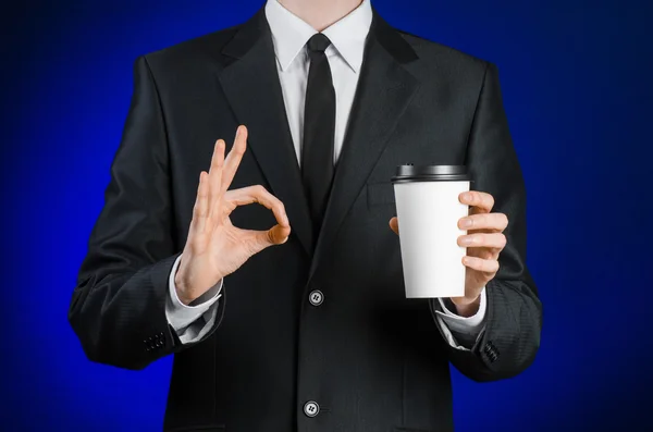 Business lunch and coffee theme: businessman in a black suit holding a white blank paper cup of coffee with a brown plastic cap on a dark blue background isolated in the studio, advertising coffee — Stok fotoğraf