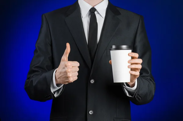 Business lunch and coffee theme: businessman in a black suit holding a white blank paper cup of coffee with a brown plastic cap on a dark blue background isolated in the studio, advertising coffee — Stockfoto