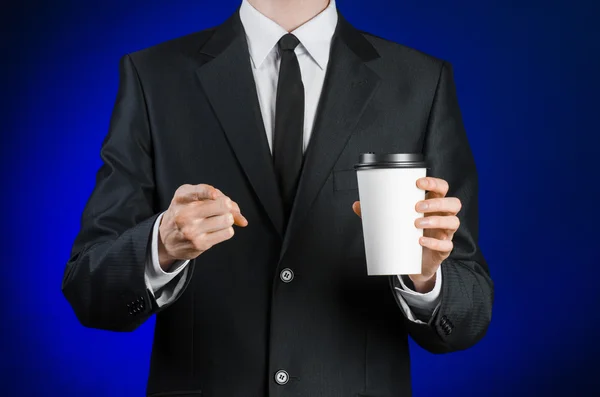 Business lunch and coffee theme: businessman in a black suit holding a white blank paper cup of coffee with a brown plastic cap on a dark blue background isolated in the studio, advertising coffee — Stockfoto