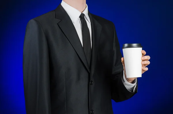 Business lunch and coffee theme: businessman in a black suit holding a white blank paper cup of coffee with a brown plastic cap on a dark blue background isolated in the studio, advertising coffee — Stock Fotó