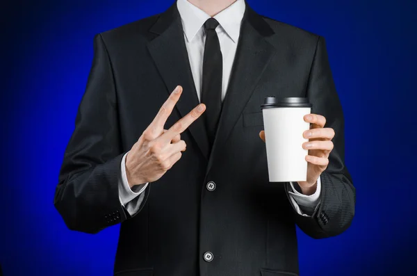 Business lunch and coffee theme: businessman in a black suit holding a white blank paper cup of coffee with a brown plastic cap on a dark blue background isolated in the studio, advertising coffee — Stock Fotó