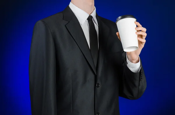 Business lunch and coffee theme: businessman in a black suit holding a white blank paper cup of coffee with a brown plastic cap on a dark blue background isolated in the studio, advertising coffee