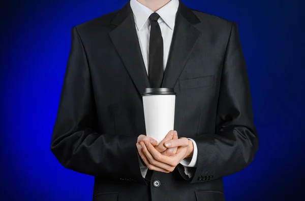 Almuerzo de negocios y café tema: hombre de negocios en un traje negro sosteniendo una taza de papel blanco de café con una tapa de plástico marrón sobre un fondo azul oscuro aislado en el estudio, publicidad de café — Foto de Stock