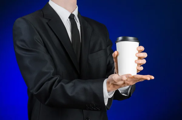 Business lunch and coffee theme: businessman in a black suit holding a white blank paper cup of coffee with a brown plastic cap on a dark blue background isolated in the studio, advertising coffee — Stok fotoğraf