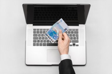 Business and freelance topic: hand in a black suit holding a banknote of 20 euro on a laptop on a white table in the studio isolated top view