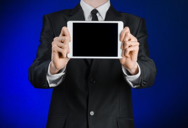 Presentation and business theme: a man in a black suit holding a white tablet touch computer gadget with touch blank black screen on a dark blue background in studio isolated