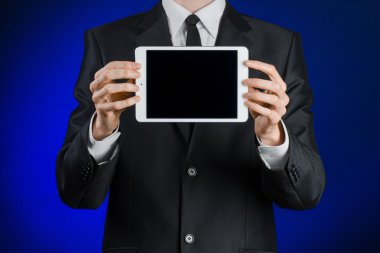 Presentation and business theme: a man in a black suit holding a white tablet touch computer gadget with touch blank black screen on a dark blue background in studio isolated