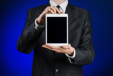 Presentation and business theme: a man in a black suit holding a white tablet touch computer gadget with touch blank black screen on a dark blue background in studio isolated