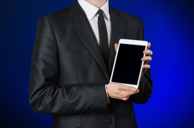 Presentation and business theme: a man in a black suit holding a white tablet touch computer gadget with touch blank black screen on a dark blue background in studio isolated