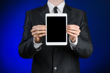 Presentation and business theme: a man in a black suit holding a white tablet touch computer gadget with touch blank black screen on a dark blue background in studio isolated