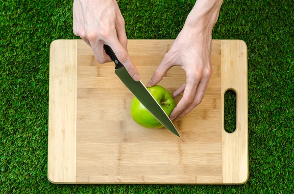 Vegetariani e cucina sulla natura del tema: mano umana che tiene un coltello e mela verde sullo sfondo di un tagliere e erba verde vista dall'alto — Foto Stock