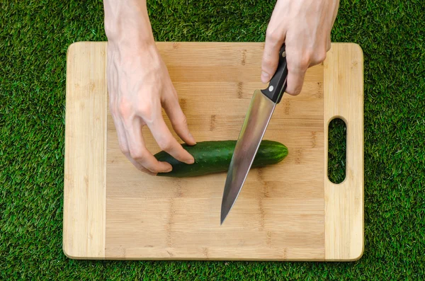 Vegetariani e cucina sulla natura del tema: mano umana che tiene un coltello e cetriolo su un tagliere e uno sfondo di erba verde vista dall'alto — Foto Stock